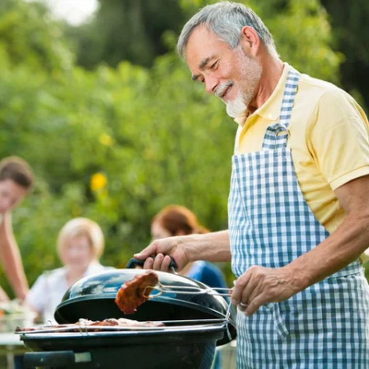 Un hombre de edad avanzada cocina en una parrilla a gas al aire libre, mientras una familia comparte en el fondo